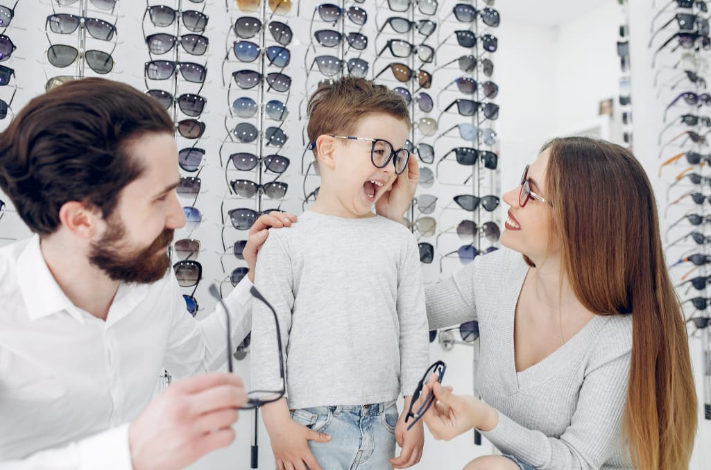 Child with parents in front of a display of glasses in an optician's shop.
