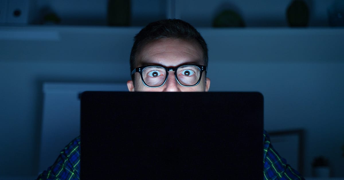 wide eyed man wearing glasses in darkened room, looking at laptop screen