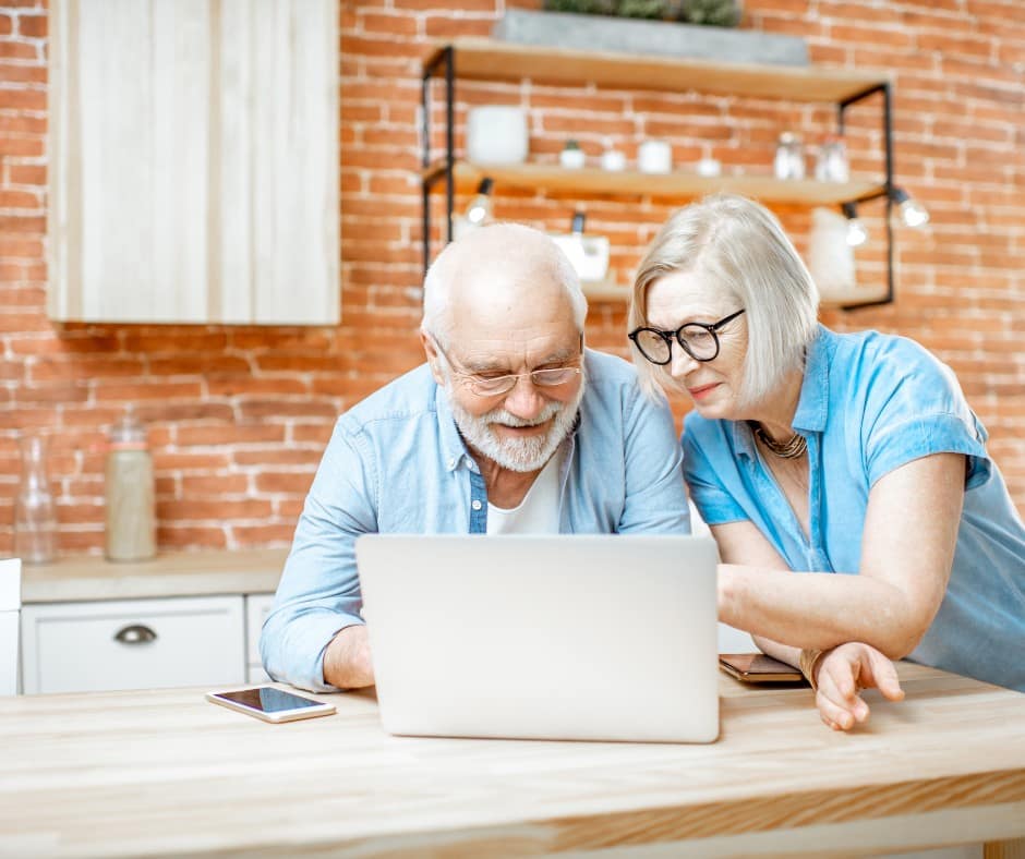 elderly couple, both wearing prescription glasses, studying a laptop screen in their kitchen